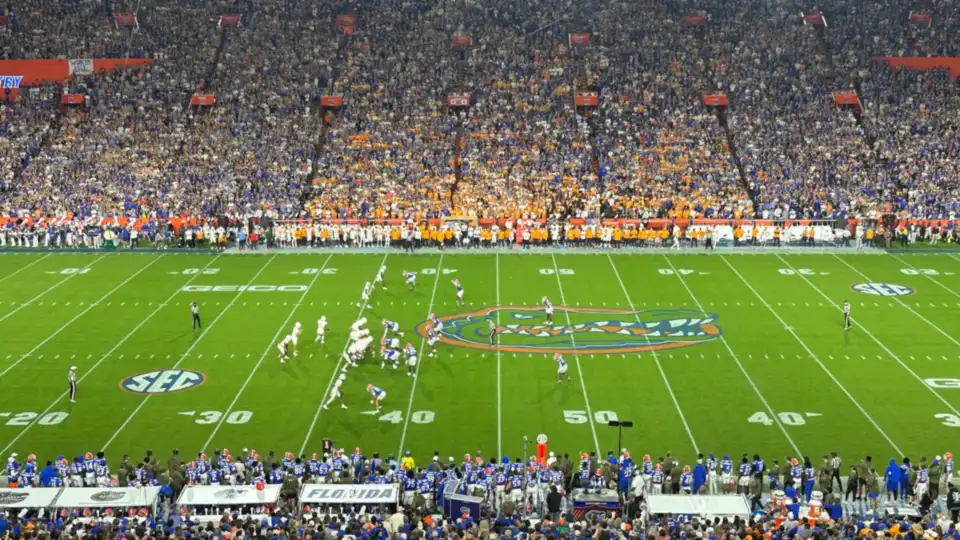 A packed college football stadium at night, seen from high in the stands, with a game in progress on the field.